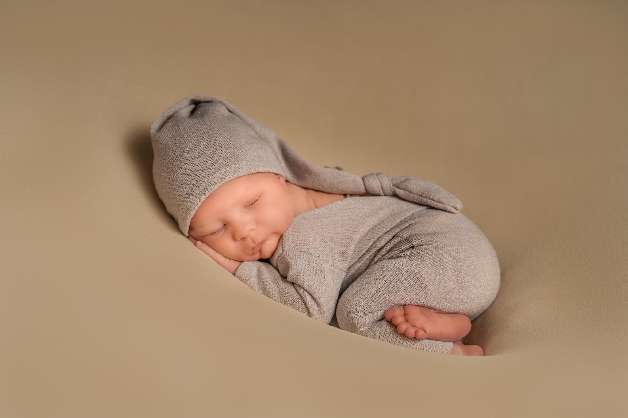 Śpiący noworodek w szarym, dzianinowym kombinezonie i czapeczce z uszkami, ułożony na beżowym tle.