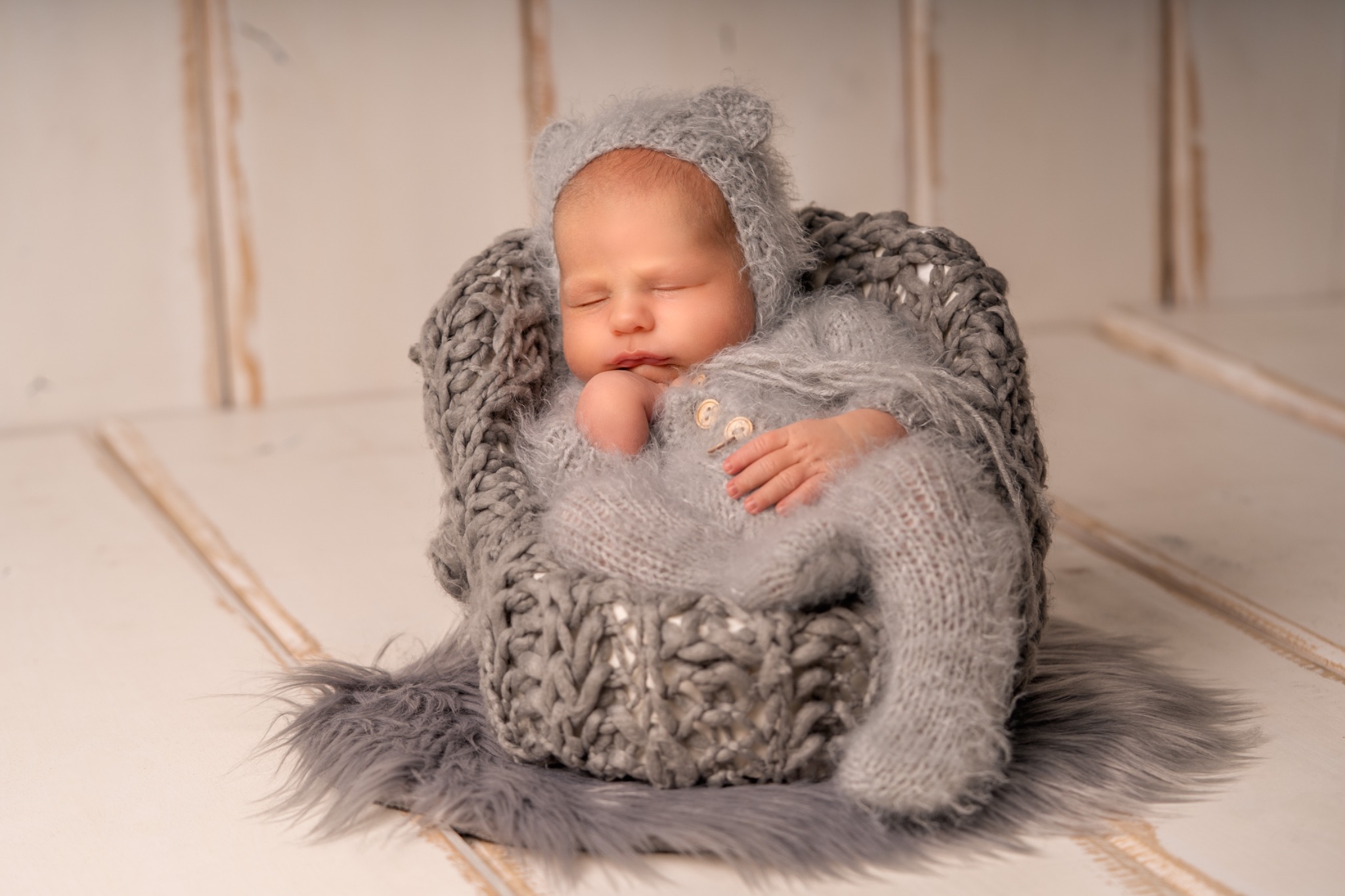 Śpiący noworodek w szarym, dzierganym kombinezonie z kapturem z uszkami, ułożony w dzierganym koszyku na futrzanym dywaniku, na drewnianym tle.