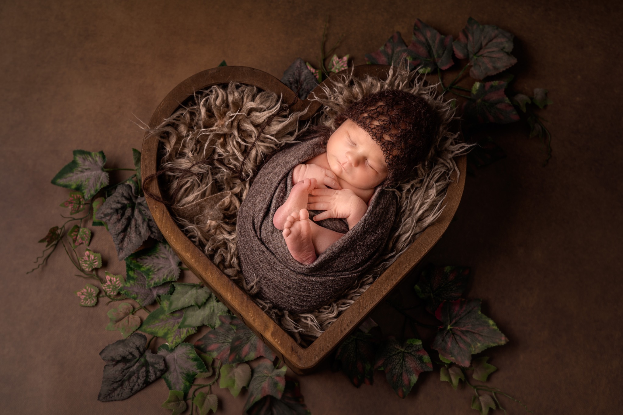 Śpiące nowo narodzone dziecko zawinięte w brązowy materiał i ułożone w drewnianym koszyku w kształcie serca, z dekoracją z liści winorośli w rogu.