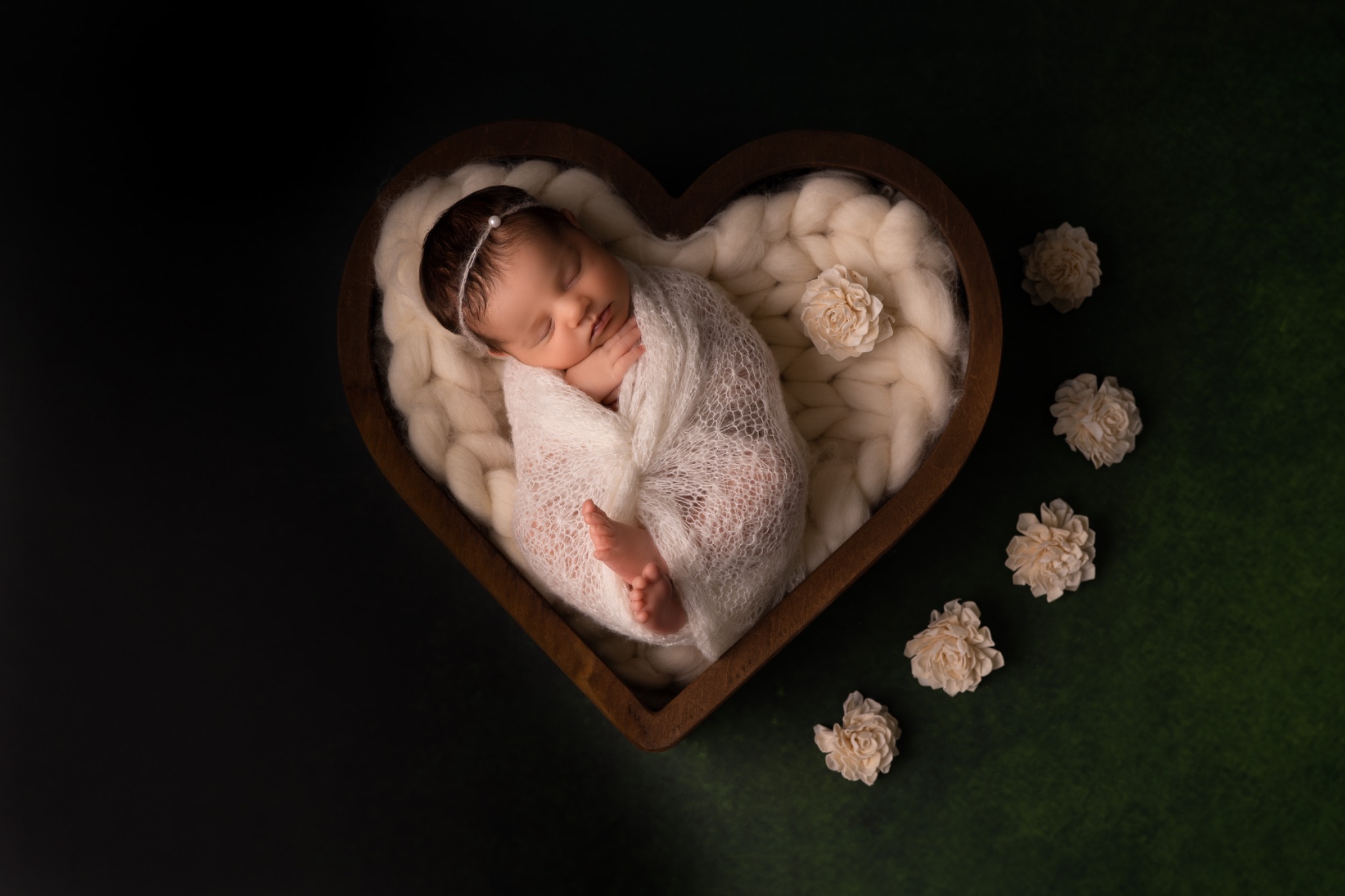 Noworodek owinięty w białą, ażurową tkaninę, śpiący w drewnianym koszyku w kształcie serca, wyłożonym puszystą wełną, z dekoracjami z kremowych kwiatów na ciemnozielonym tle.
