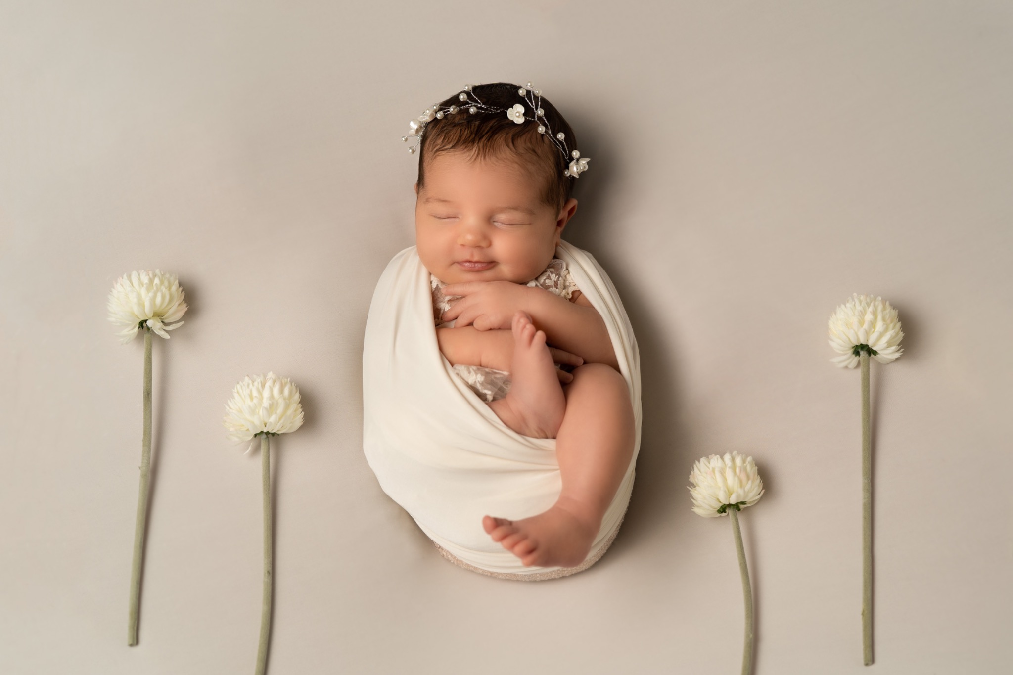 Śpiące niemowlę zawinięte w kremowy materiał, ozdobione wiankiem z perełek i kwiatów, otoczone czterema białymi kwiatami na gładkim, jasnym tle.