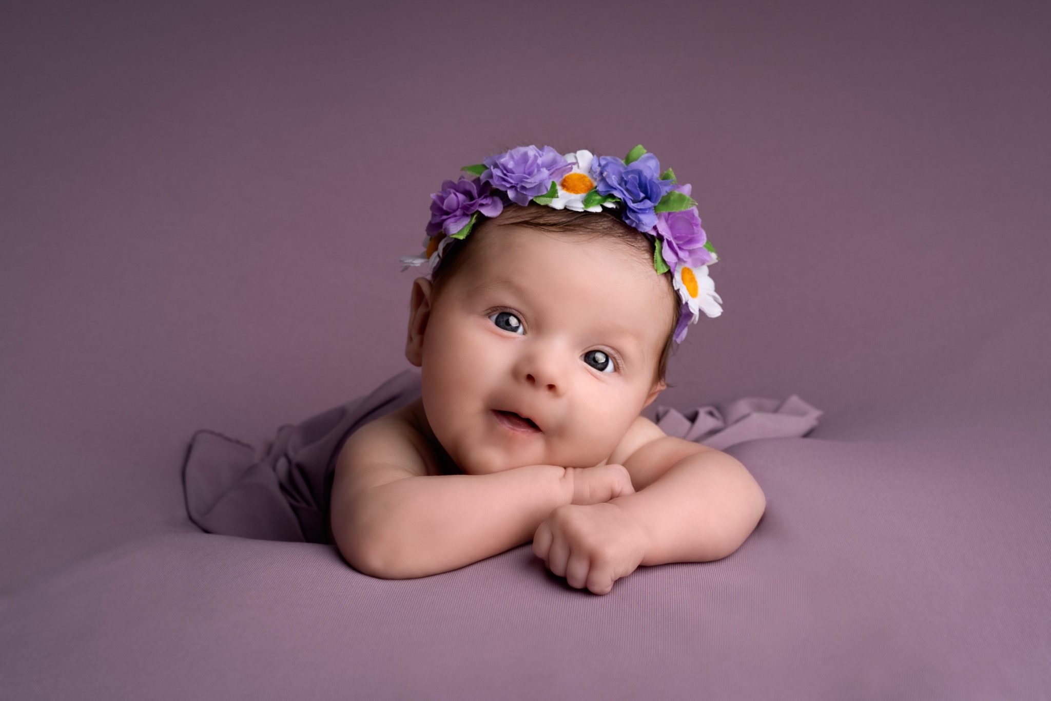Uśmiechnięty noworodek leży na fioletowym tle, ubrany w wianek z fioletowych, białych i niebieskich kwiatów.