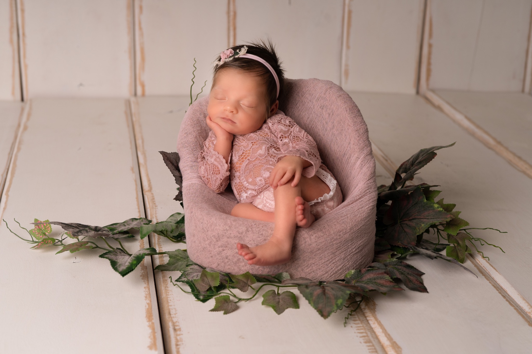 Śpiąca noworodka w koronkowej kreacji, ułożona w różowym foteliku otoczonym dekoracyjnymi liśćmi, na tle białych, drewnianych desek.