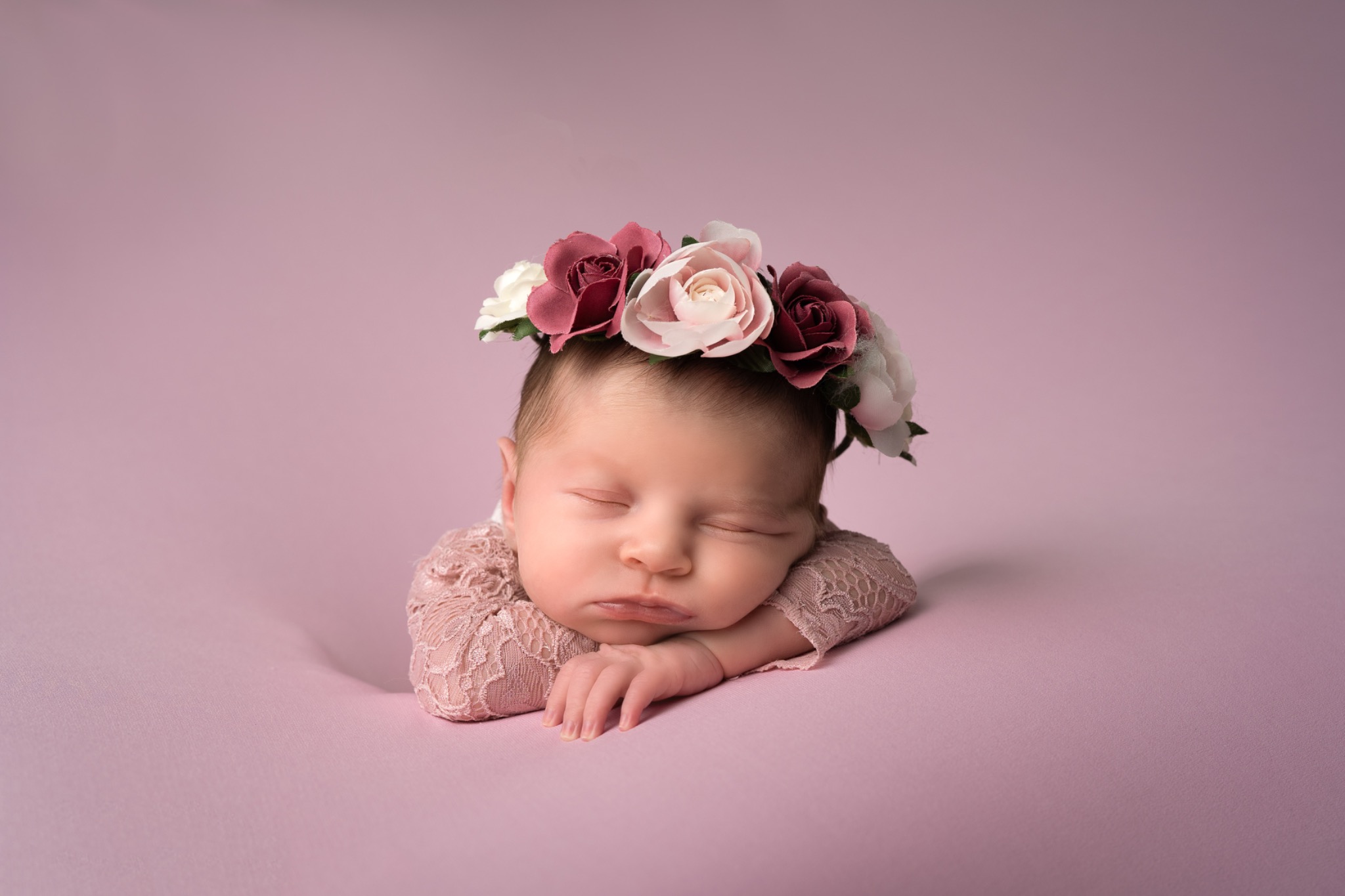 Śpiące nowonarodzone dziecko w koronie z różowych i białych róż, ubrane w koronkową sukienkę, na różowym tle.