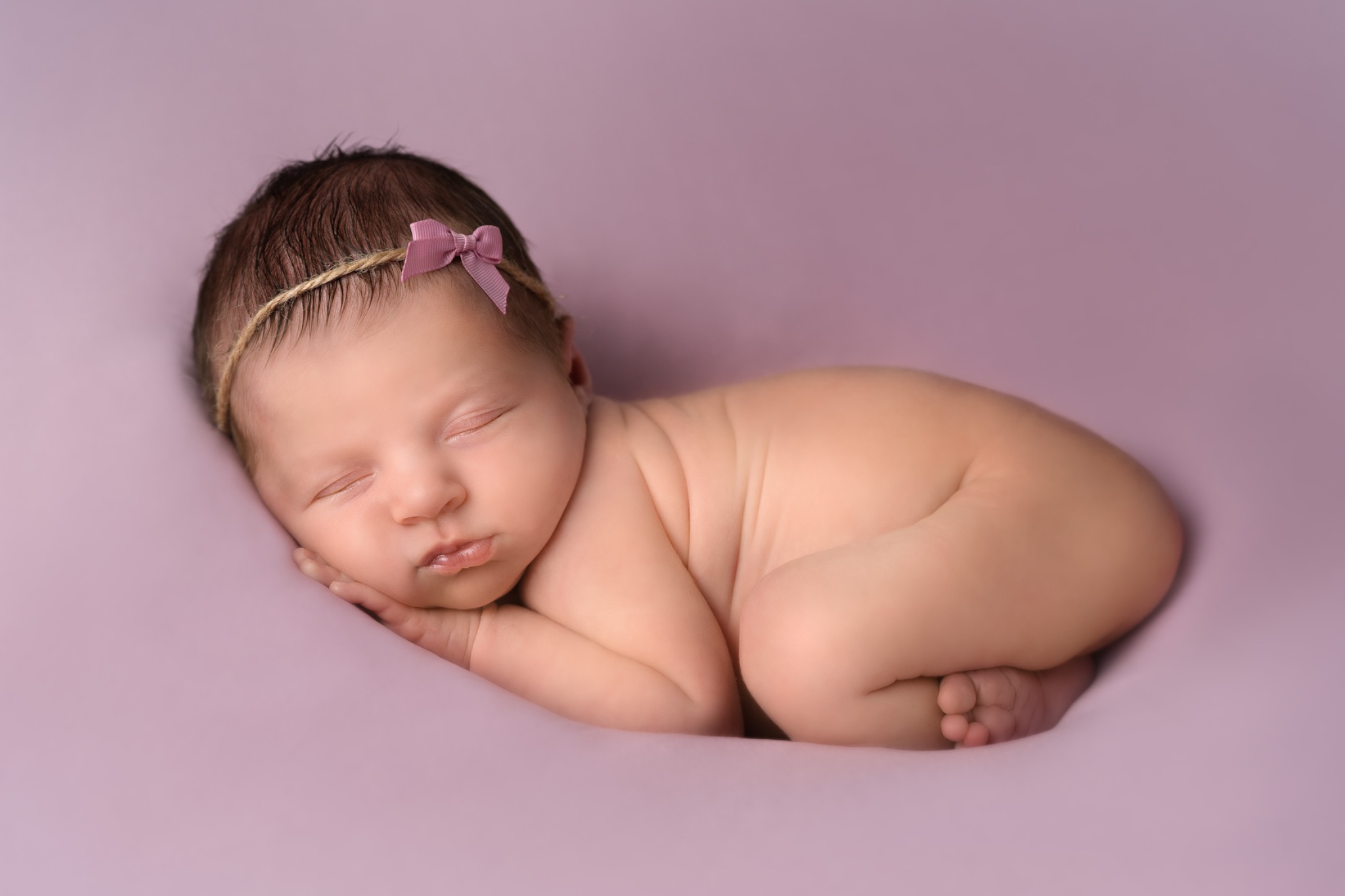 Śpiący noworodek z ciemnymi włosami, owinięty w kokon na fioletowym tle, z delikatną opaską z kokardką na głowie.