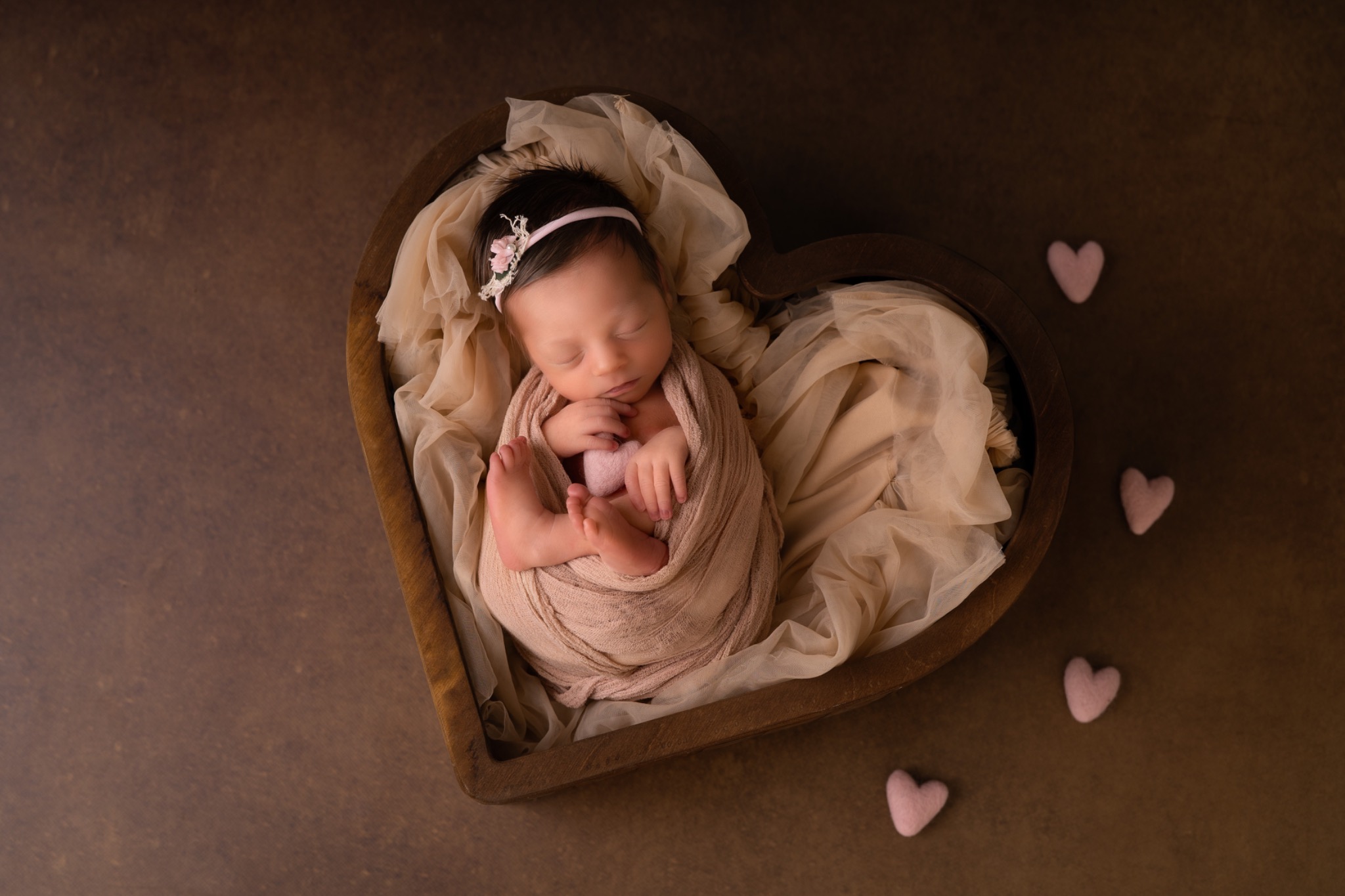 Noworodek zawinięty w beżowy materiał, śpiący w drewnianej misce w kształcie serca, z małą opaską z kwiatkiem na głowie i trzema filcowymi sercami obok.
