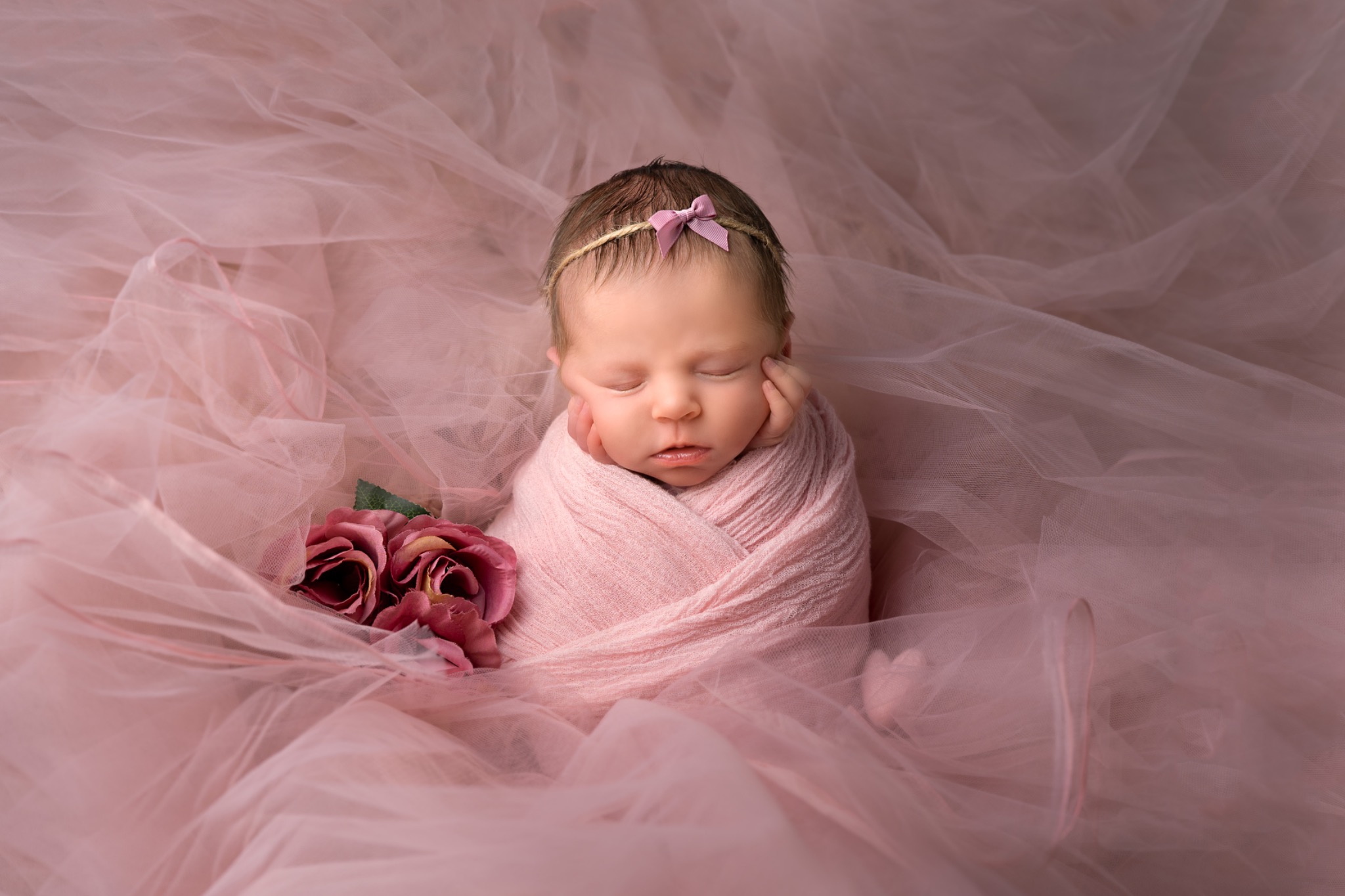 Śpiące nowonarodzone dziecko owinięte w różowy materiał, z różową opaską z kokardką na głowie, leżące na tle z różowego tiulu i różowych kwiatów.