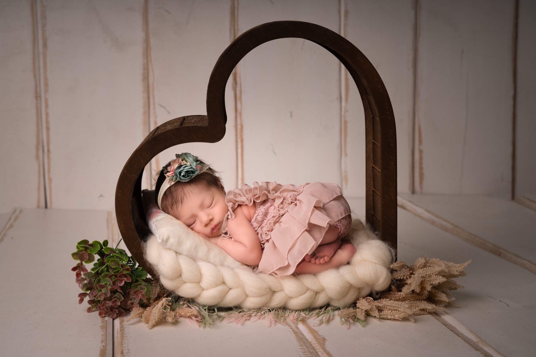 Śpiąca noworodka w różowej sukience i z kwiatową opaską na głowie, ułożona w drewnianej ramce w kształcie serca na puszystym, kremowym kocyku z dodatkiem zielonych gałązek.