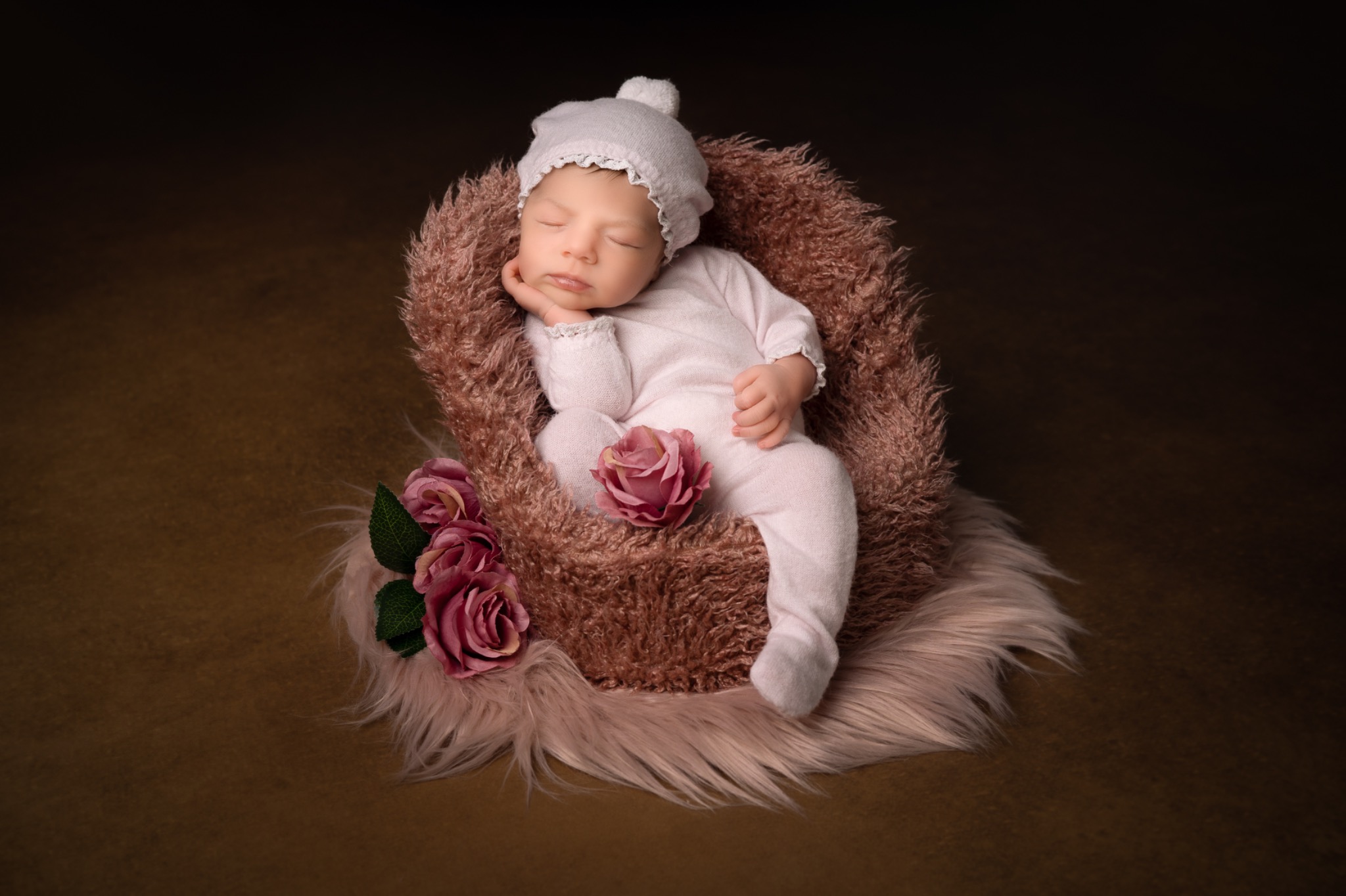 Śpiący noworodek w jasnoróżowym ubranku i czapeczce, ułożony w brązowym futerkowym koszyku z dekoracją z różowych róż.