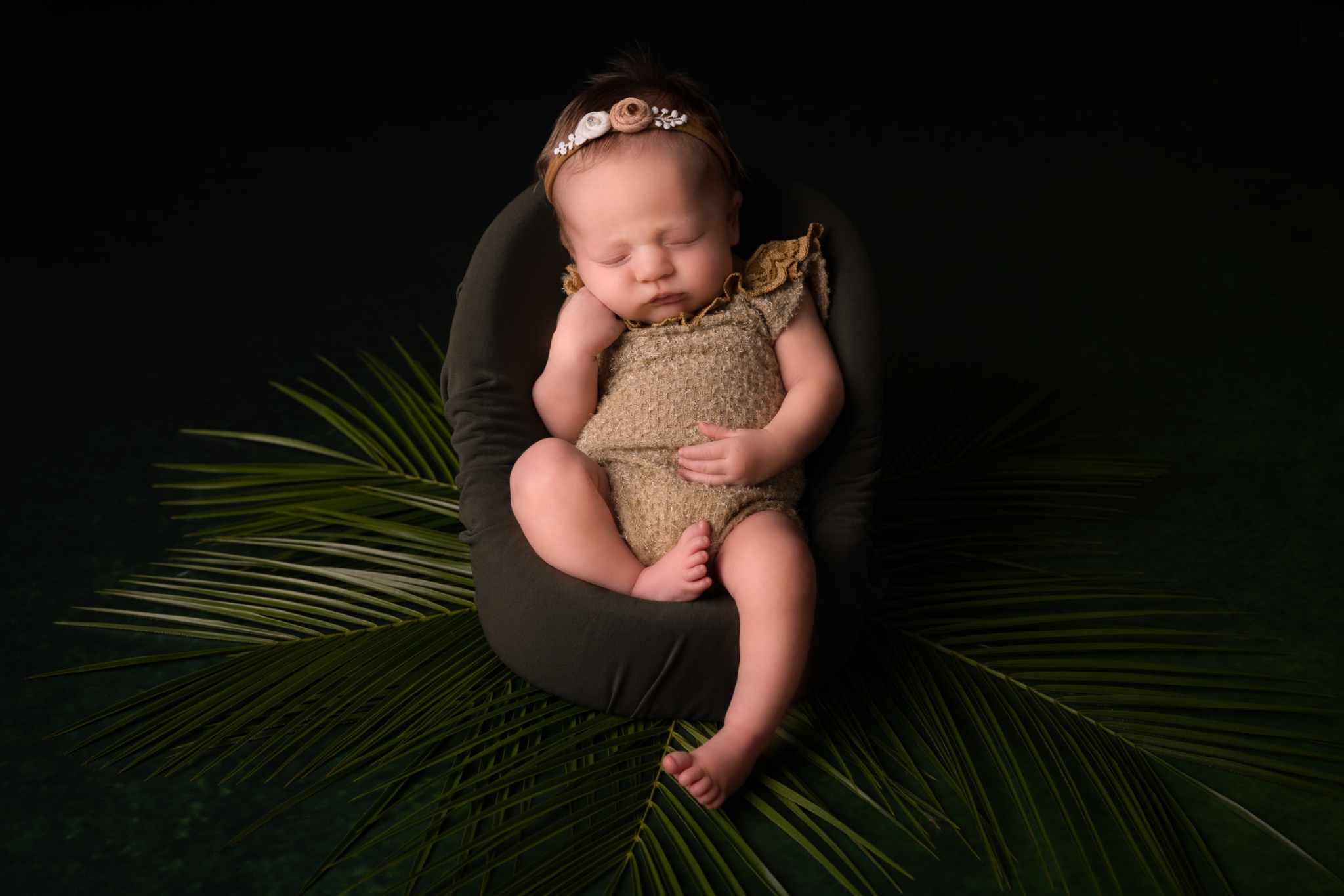 Śpiący noworodek w beżowym ubranku, z opaską z kwiatami na głowie, ułożony w ciemnozielonym fotelu na tle liści palmy.