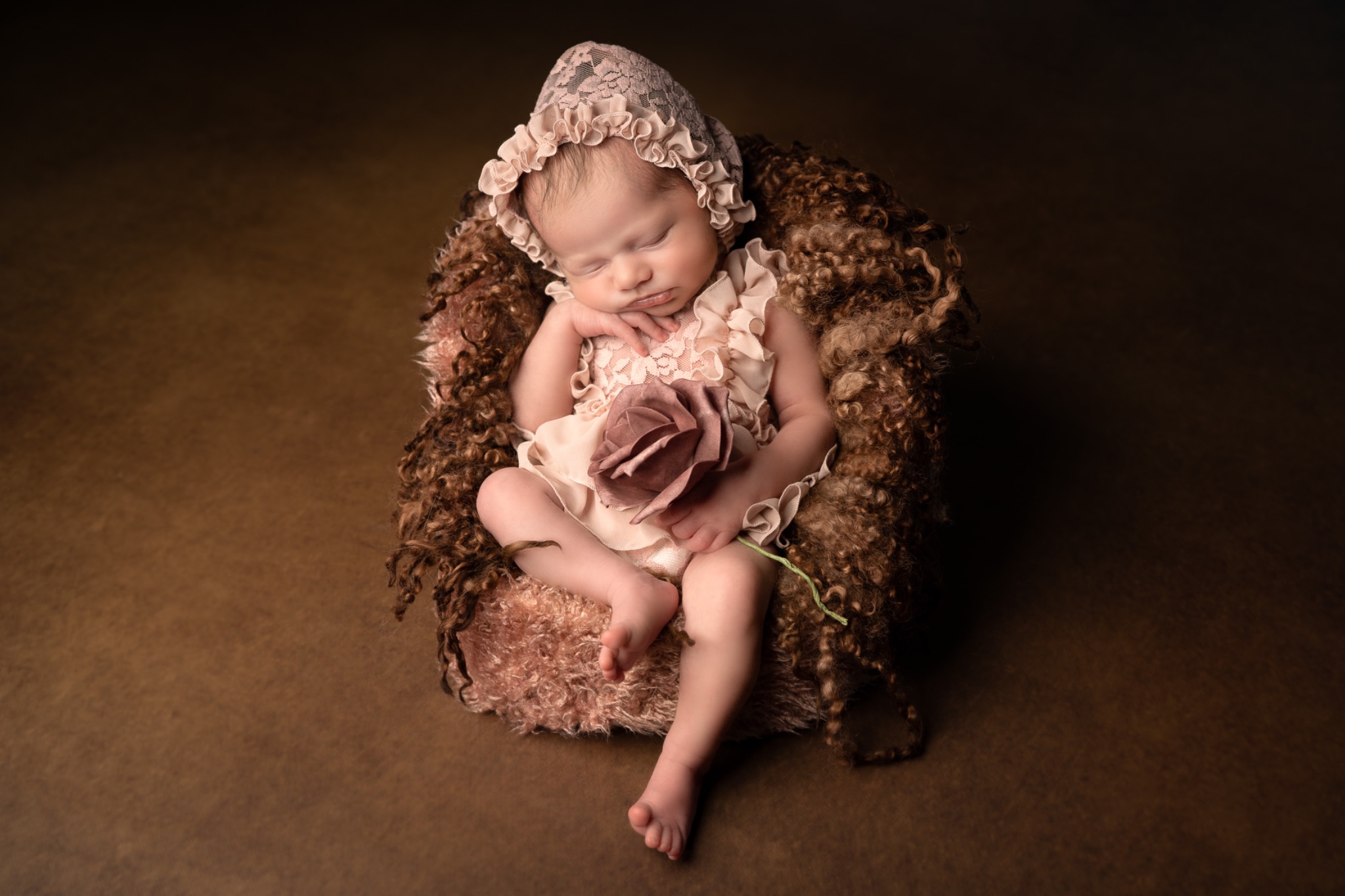 Śpiąca noworodka w koronkowej czapeczce, ułożona w brązowym, teksturowanym gnieździe z dekoracyjnym kwiatem, na jednolitym, brązowym tle.