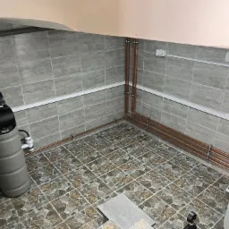 Szara stacja uzdatniania wody Aitech z manometrami, miedziane rury instalacji wodnej w pomieszczeniu wyłożonym szarą, drewnopodobną płytką ceramiczną.