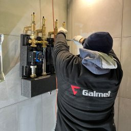 Fach-Mach Paweł Kalisz - Instalator w czarnej bluzie z logo Galmet montuje pompę ciepła w pomieszczeniu z szarymi płytkami, używając śrubokręta.