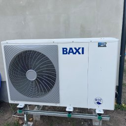 Fach-Mach Paweł Kalisz - Zewnętrzna jednostka klimatyzacji marki BAXI, zamontowana na metalowej konstrukcji przy szarej ścianie budynku, z widocznym niebieskim panelem izolacyjnym obok i osobą w tle.