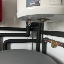 Fach-Mach Paweł Kalisz - Wnętrze kotłowni: biały zasobnik ciepłej wody Galmet z widocznymi czarnymi rurami w izolacji termicznej, armatura z czerwonymi zaworami i naklejka serwisu na zbiorniku.