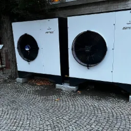 Dwie białe jednostki zewnętrzne pompy ciepła Galmet Airmax2 z czarnymi wentylatorami, umieszczone na betonowych podstawach przed budynkiem z cegły, obok drzewa na brukowanej powierzchni.