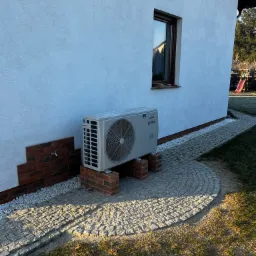 Zewnętrzna jednostka klimatyzacji typu split, model Prima, zamontowana na ceglanych postumentach przy białej ścianie domu z widocznym oknem i brukowanym chodnikiem w słoneczny dzień.