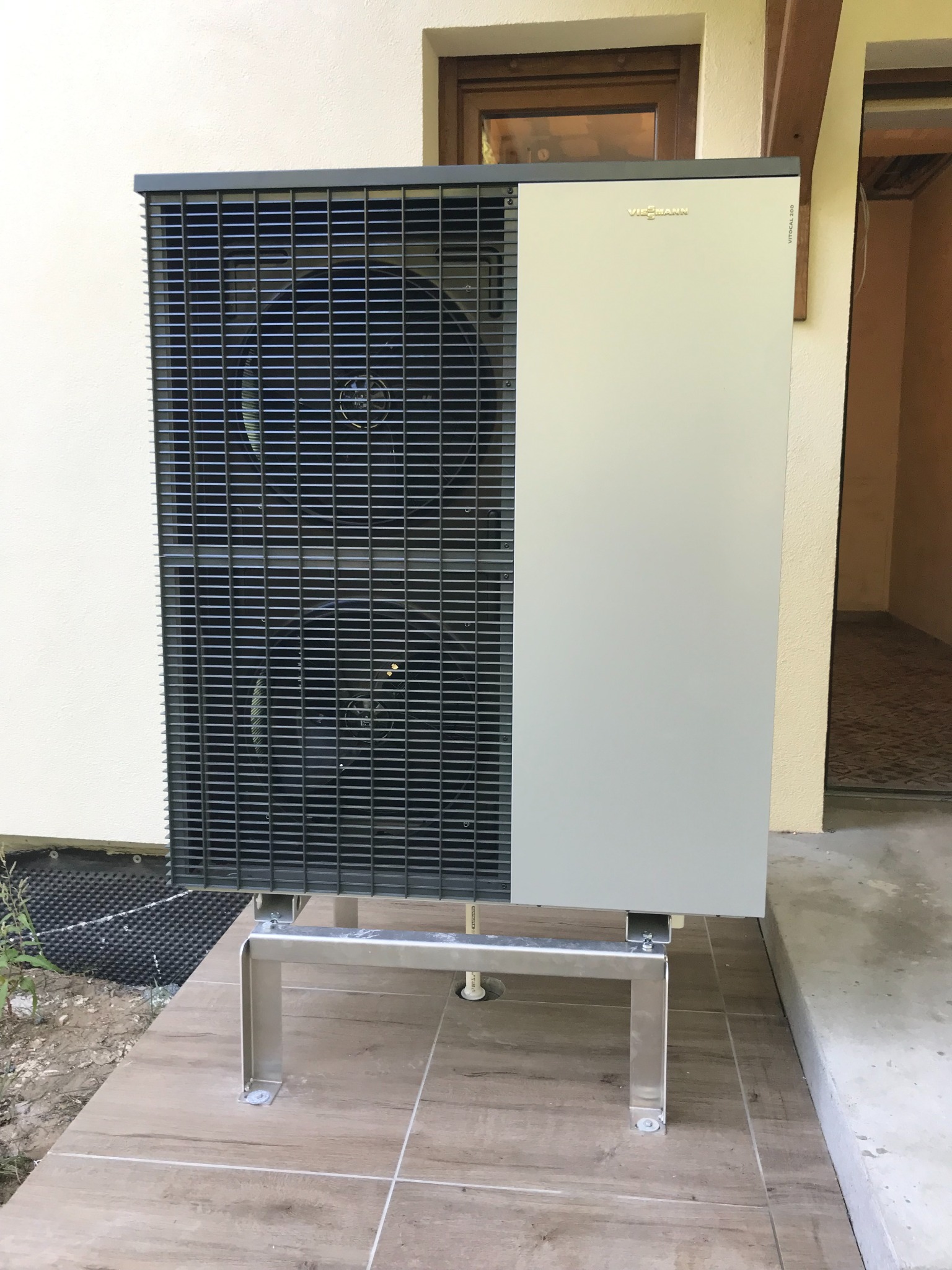 Zewnętrzna jednostka pompy ciepła Viessmann Vitocal 200, zamontowana na metalowym stelażu na płytkach imitujących drewno, widoczna przy ścianie budynku z oknem.