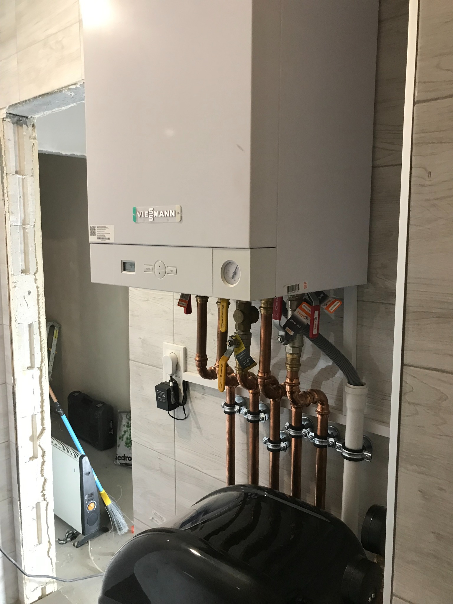 Biały piec grzewczy Viessmann zamontowany na ścianie z beżowych płytek, widoczne miedziane rury, zawory i podłączenia, w tle niedokończone wykończenie ściany i podłogi.