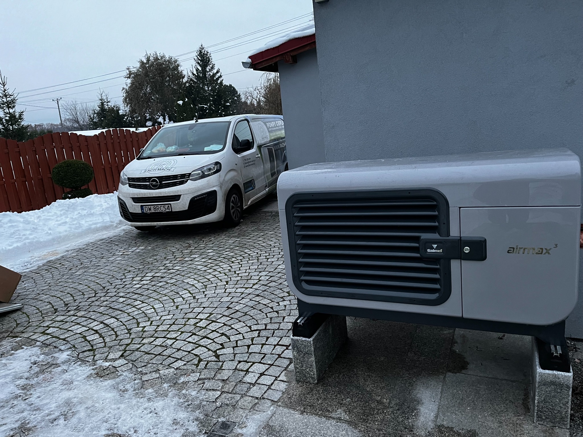 Szara pompa ciepła Airmax³ zamontowana na betonowych bloczkach przed szarą ścianą budynku, z firmowym vanem z logo Fach-Mach zaparkowanym na kostce brukowej w zimowej scenerii.