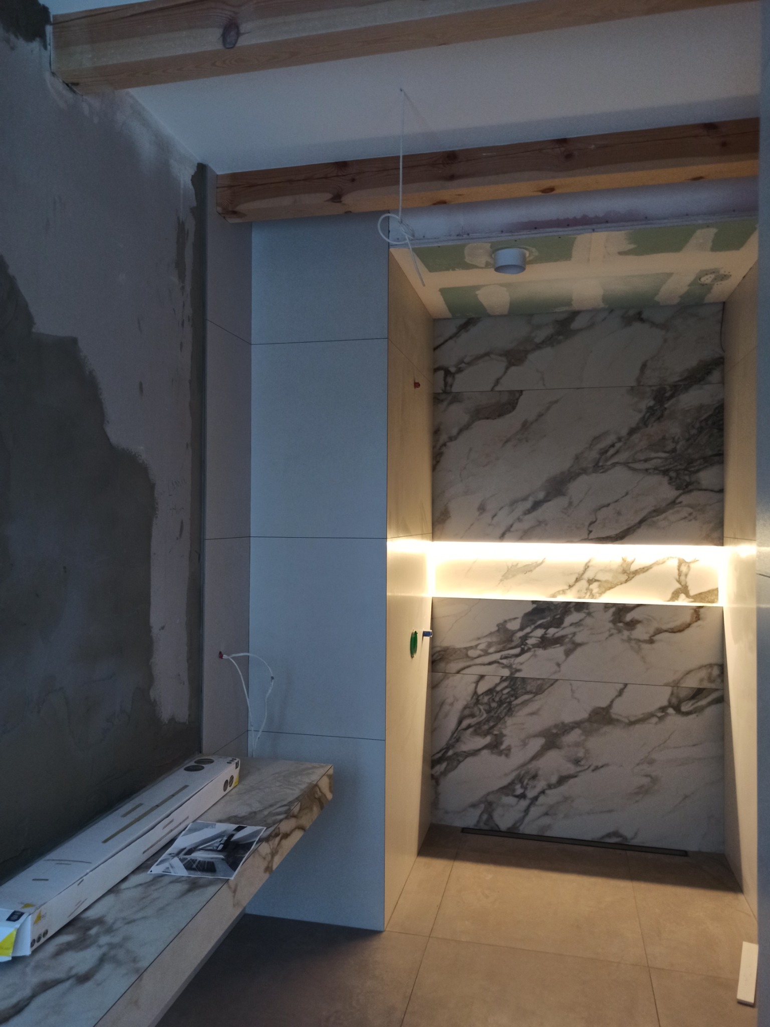 Wnęka prysznicowa w trakcie wykańczania, z podświetleniem LED między panelami imitującymi marmur, widoczne niedokończone ściany i sufit z elementami drewnianymi.