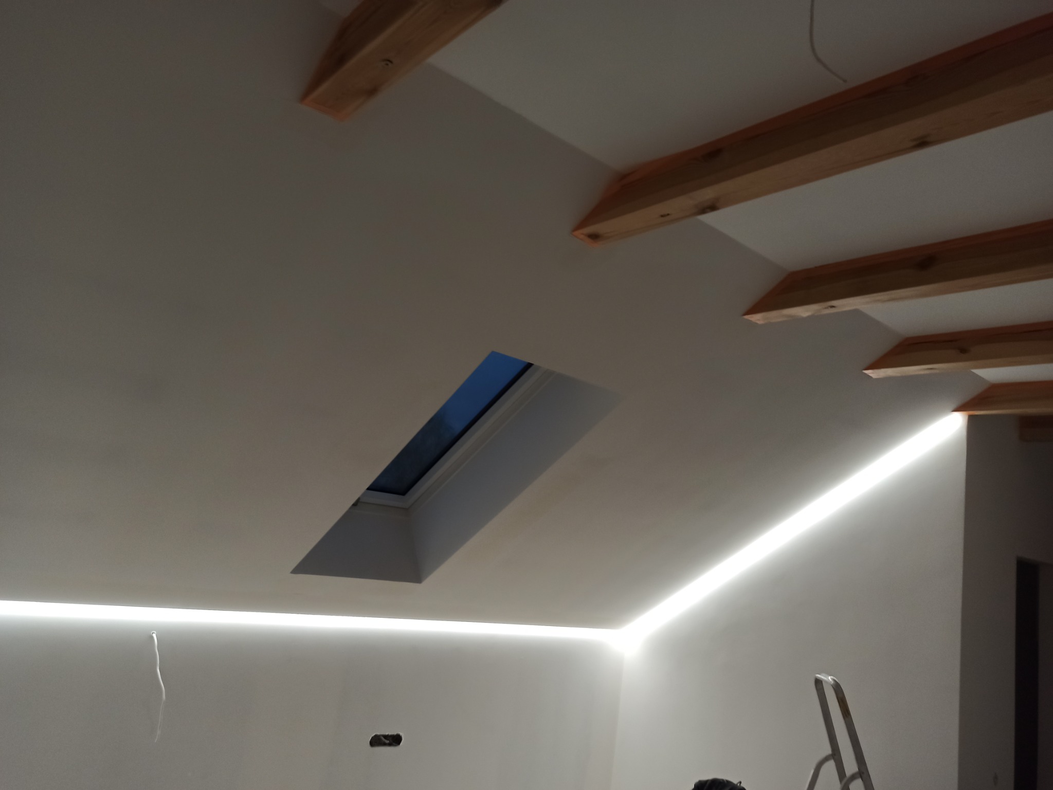 Poddasze z oknem dachowym i ukrytym liniowym oświetleniem LED wzdłuż krawędzi sufitu, widoczne elementy drewniane konstrukcji dachu i drabina malarska.