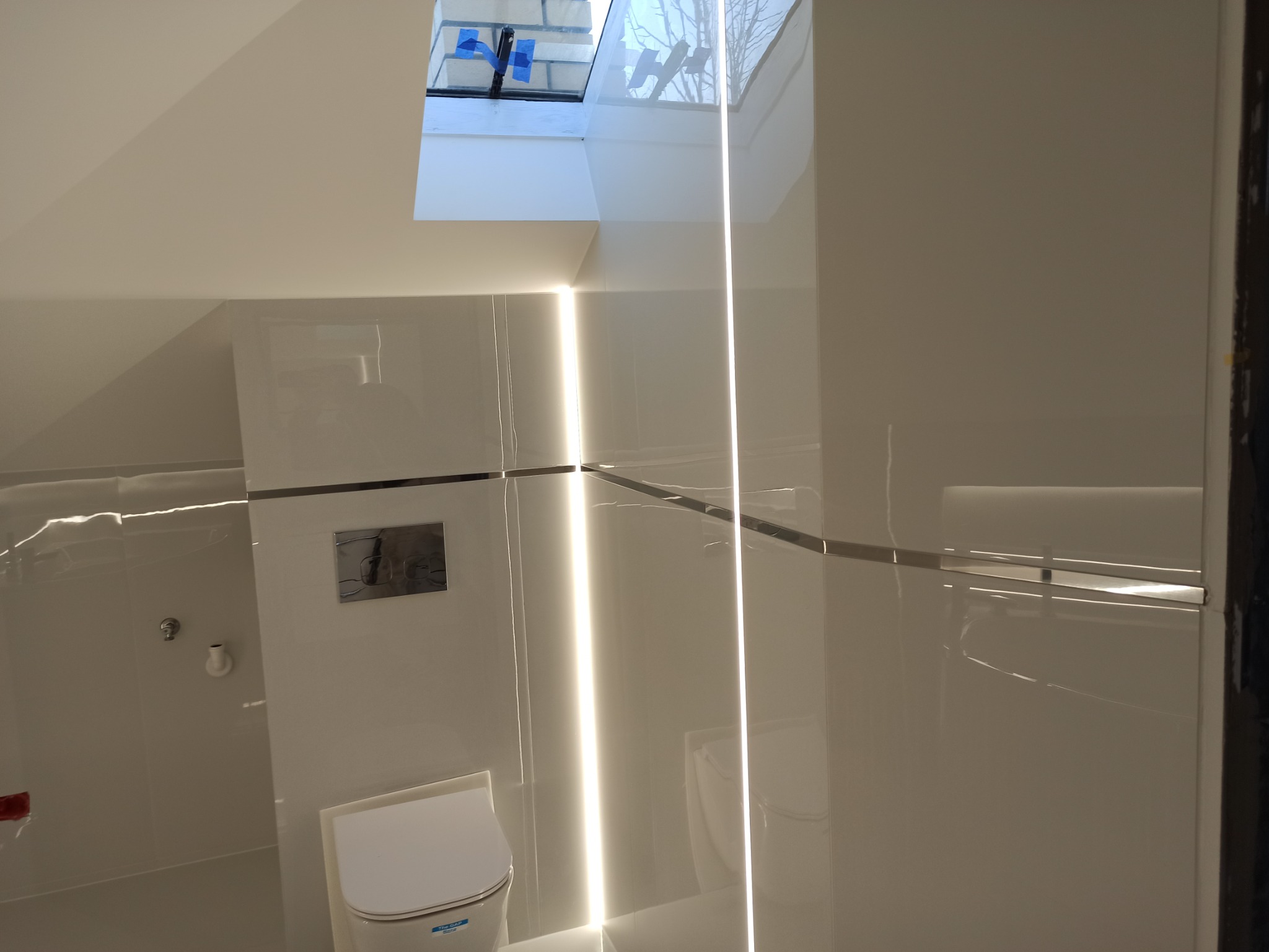 Nowoczesna, biała łazienka z podświetlanymi panelami ściennymi i oknem dachowym, ukazująca minimalistyczny design i czystość form.