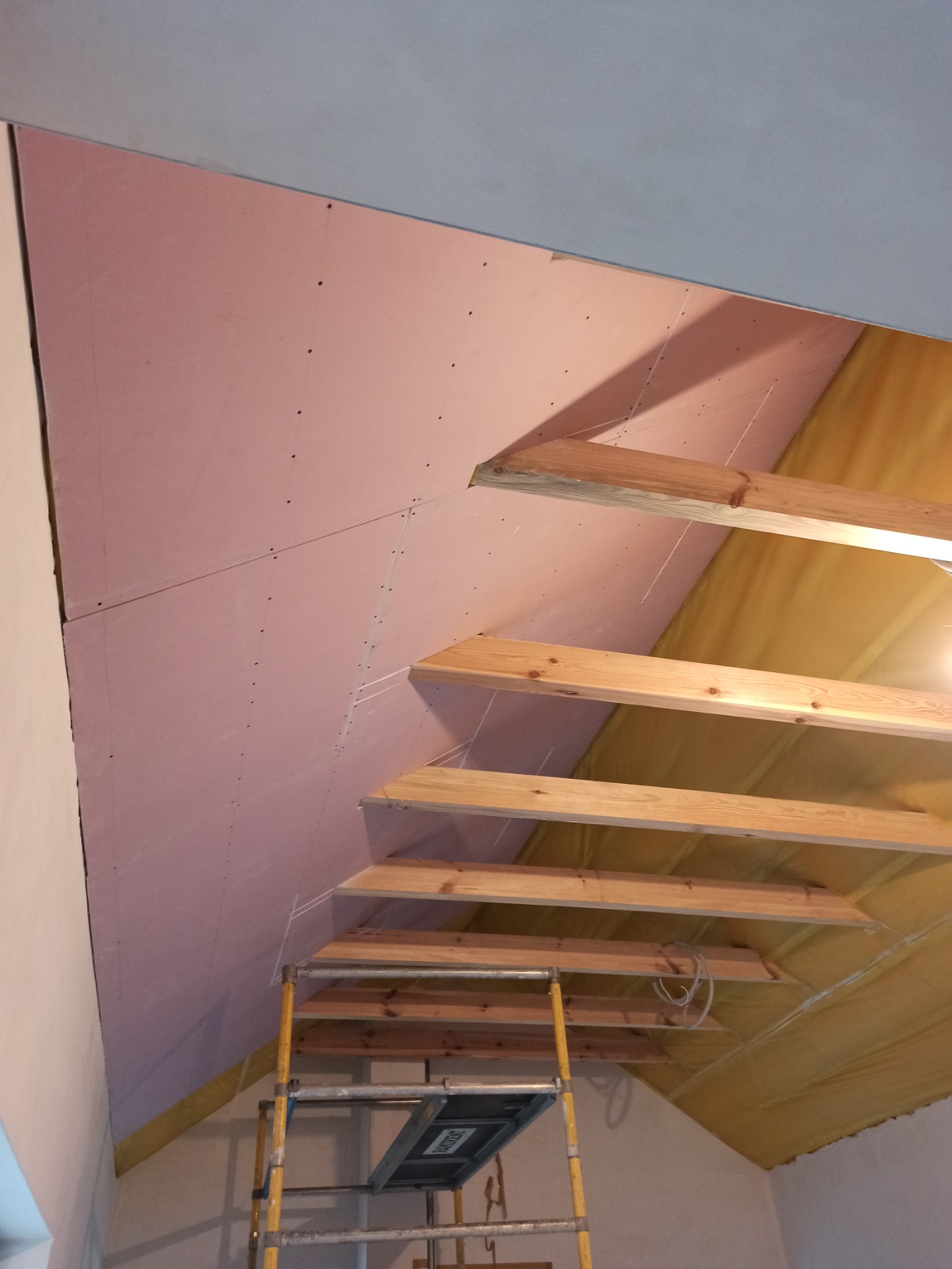 Montaż różowej płyty karton gips na skośnym poddaszu, widoczne drewniane belki konstrukcyjne i drabina monterska.