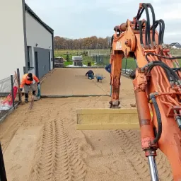 Przygotowanie podłoża pod kostkę brukową w Strzelcach Opolskich: robotnicy wyr&oacute;wnują teren piaskiem, widoczna koparka Atlas i ułożone obok kostki brukowe.