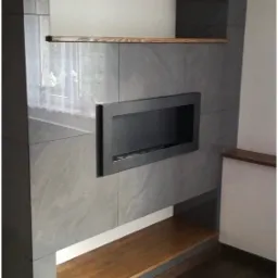 Szara ściana z płytek imitujących kamień, z wnęką na kominek i drewnianymi półkami, na podłodze ciemne panele.