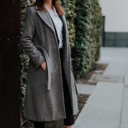 Kobieta w szarym płaszczu z ręką w kieszeni, stojąca na chodniku obok ściany z bluszczem, ubrana w białą koszulę i czarne spodnie, szare szpilki.