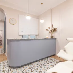 Nowoczesne wnętrze biura z szarą ladą recepcyjną, krzesłami z białej skóry i drewnianymi elementami, oraz wzorzystą podłogą w odcieniach beżu i błękitu.