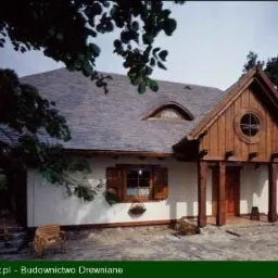 Gotowy dom szkieletowy z drewnianą fasadą, okrągłym oknem na szczycie i małym oknem na poddaszu, otoczony zielenią.