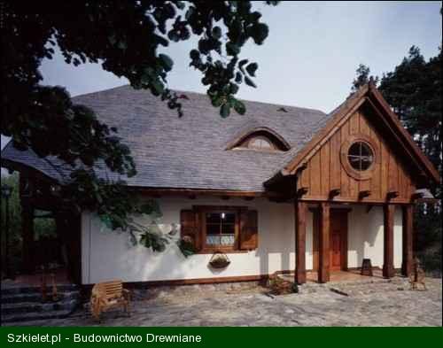 Gotowy dom szkieletowy z drewnianą fasadą, okrągłym oknem na szczycie i małym oknem na poddaszu, otoczony zielenią.