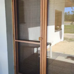 Drewniane drzwi balkonowe z zamontowaną moskitierą w drewnianej ramie, widoczne żaluzje wewnętrzne, z zewnątrz widać fragment elewacji i kostki brukowej z wycieraczką.