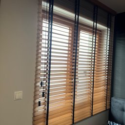 Okna LaVico Piotr Wontorczyk - Zamknięte drewniane żaluzje z czarnymi taśmami na oknie, widoczne fragmenty ściany i kanapy.