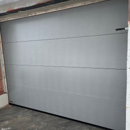 Okna LaVico Piotr Wontorczyk - Zamknięta, segmentowa brama garażowa w kolorze szarym, widoczna podczas montażu w surowym otoczeniu budowy, z nożem wbitą w ocieplenie ściany obok.