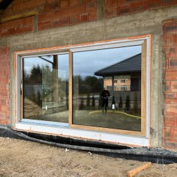 Okna LaVico Piotr Wontorczyk - Montaż dużego okna przesuwnego w surowym stanie, widoczna pianka montażowa, ceglany mur i odbicie budynku w szybie.