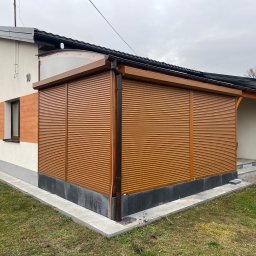 Okna LaVico Piotr Wontorczyk - Zabudowa tarasu brązowymi roletami zewnętrznymi z widocznym systemem prowadnic i skrzynką górną, zadaszenie z poliwęglanu, elewacja budynku z elementami drewnopodobnymi.