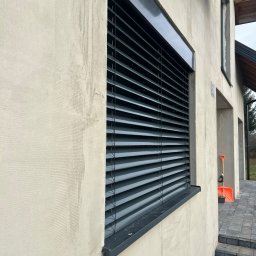 Okna LaVico Piotr Wontorczyk - Szare żaluzje zewnętrzne zamontowane na elewacji budynku z widocznym oknem obok i schodami wejściowymi.