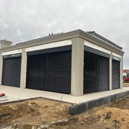 Okna LaVico Piotr Wontorczyk - Nowoczesny budynek z szarym tynkiem i czarnymi, zewnętrznymi roletami, w trakcie prac wykończeniowych na zewnątrz, widoczne rury kanalizacyjne i rozkopany teren.