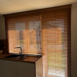 Okna LaVico Piotr Wontorczyk - Zamontowane żaluzje drewniane w kuchni w Niechcicach, widok z wnętrza na okno z częściowo zasłoniętymi lamelami, przez które prześwituje zieleń ogrodu.