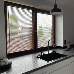 Okna LaVico Piotr Wontorczyk - Kuchnia z widokiem na zewnątrz przez okno z drewnianymi żaluzjami, na blacie czarny zlew i bateria.