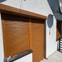 Brązowe rolety zewnętrzne zamontowane na elewacji domu z widoczną anteną satelitarną, uchwycone w słoneczny dzień.
