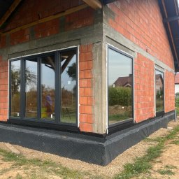 Okna LaVico Piotr Wontorczyk - Surowy stan budynku z czerwonej cegły z zamontowanymi ciemnoszarymi oknami. Widoczna czarna folia fundamentowa i częściowo wykończona elewacja.