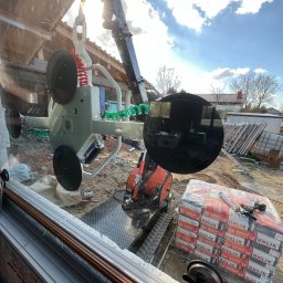 Okna LaVico Piotr Wontorczyk - Montaż okna za pomocą specjalistycznego podnośnika próżniowego. W tle widoczne materiały budowlane i fragment konstrukcji budynku w trakcie prac wykończeniowych.