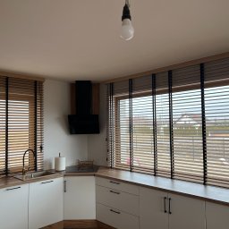 Okna LaVico Piotr Wontorczyk - Kuchnia z drewnianymi żaluzjami w oknach, jasne szafki i drewniany blat. Widok na zewnątrz przez okno. Minimalistyczny design.