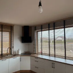 Kuchnia z drewnianymi żaluzjami w oknach, jasne szafki i drewniany blat. Widok na zewnątrz przez okno. Minimalistyczny design.