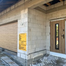 Okna LaVico Piotr Wontorczyk - Brama garażowa segmentowa z drewnianym panelem i drzwi wejściowe z szybą, zamontowane w surowym budynku z bloczków. Tablica informacyjna budowy.