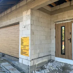 Brama garażowa segmentowa z drewnianym panelem i drzwi wejściowe z szybą, zamontowane w surowym budynku z bloczków. Tablica informacyjna budowy.